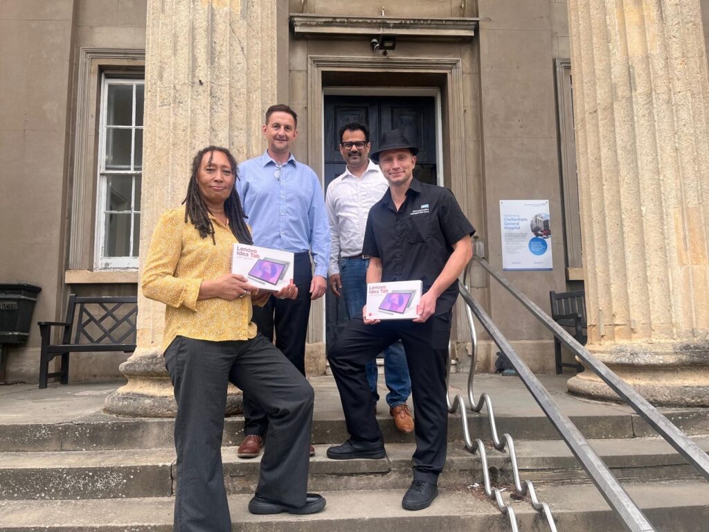 Four members of the EMOS project team stand on the steps outside Cheltenham General Hospital, holding Lenovo tablets used for the new Electronic Meal Ordering System (EMOS) rollout. The group is positioned in front of the hospital's columned entrance to celebrate the launch of the digital meal ordering system.